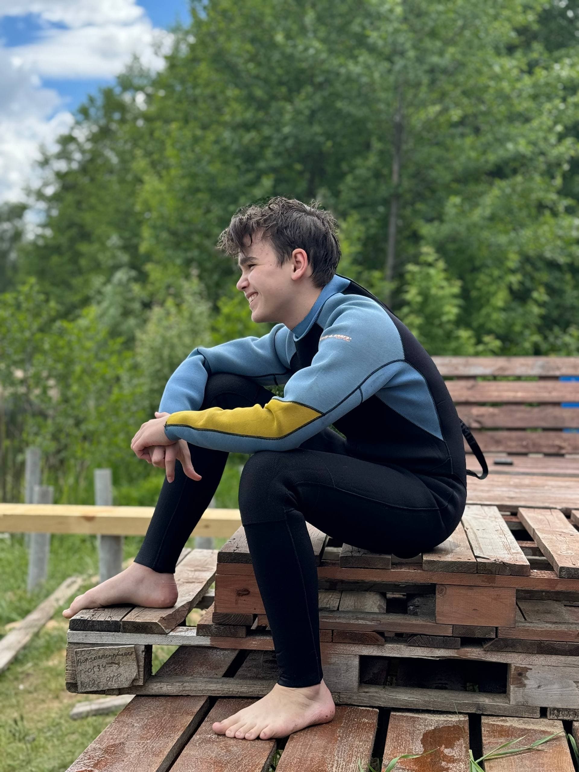 Подросток в гидрокостюме отдыхает на деревянном настиле у воды — школа Wake Park RIDE & VIBE
